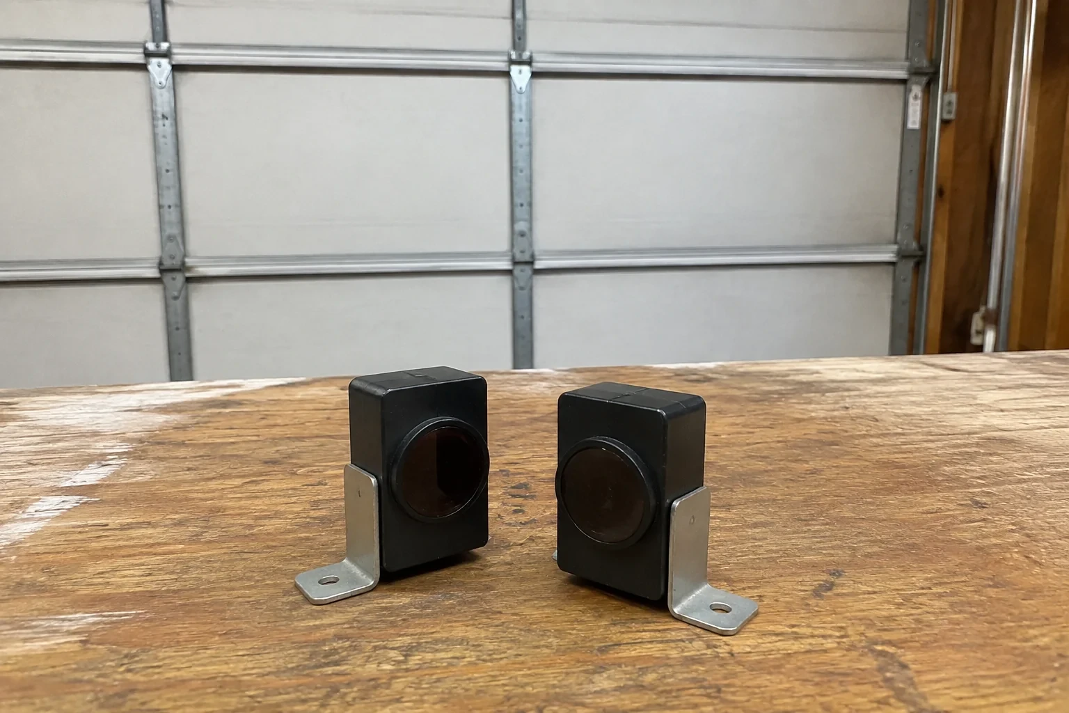 Two detached black sensor housings with round lenses and metal brackets.