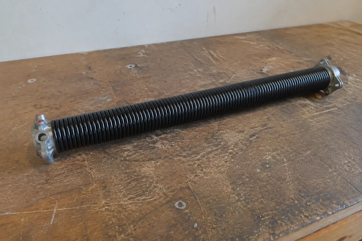 Detached black coil spring with metal end fittings on a workbench.